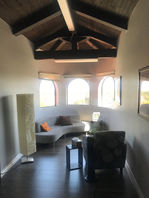 photo Woodland Hills Office Space, large white curved sofa with pillows, brown chair, tall cream colored fabic lamp, three bay windows, beamed ceiling